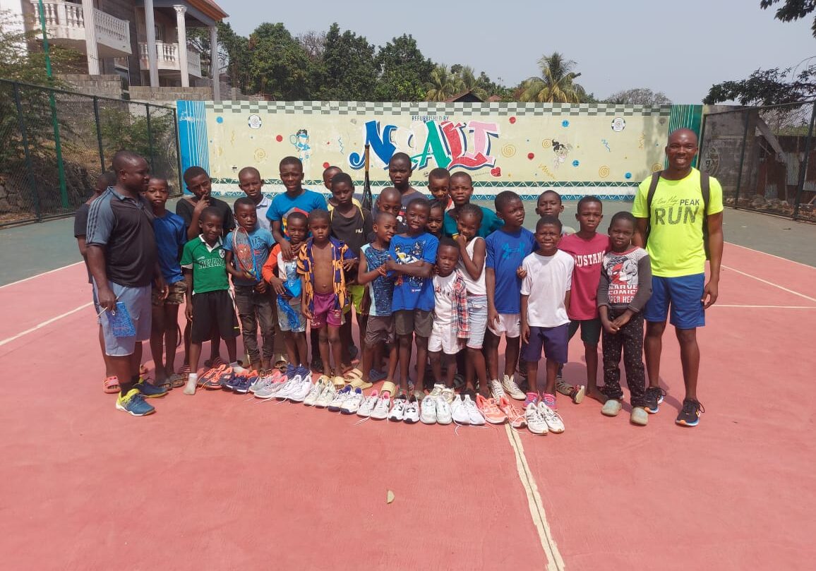 Children holding new tennis shoes in front of the court wall